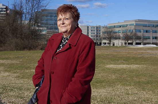 Mary Gwyn, 64, a woman with short red hair wearing a red winter coat, stands smiling in a grassy area with an office building in the background.